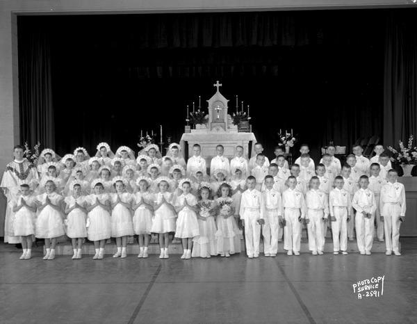 Group portrait of first communion group standing in front of the altar with a priest, at Immaculate Heart of Mary Catholic Church, 5105 Schofield Avenue.