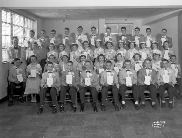 Indoor group portrait of boys and girls, in school uniforms, at graduation from a Catholic School. A priest is standing on the left.