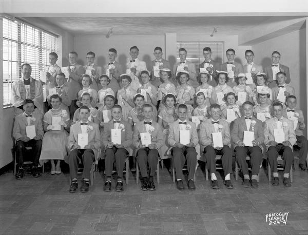 Indoor hroup portrait of boys and girls, in school uniforms, at graduation from a Catholic School. A priest is standing on the left.