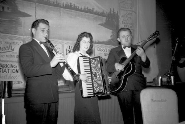 Three musicians, from left: male clarinetist, female accordion player and male guitarist standing in front of printed stage screen at the Marine Club.