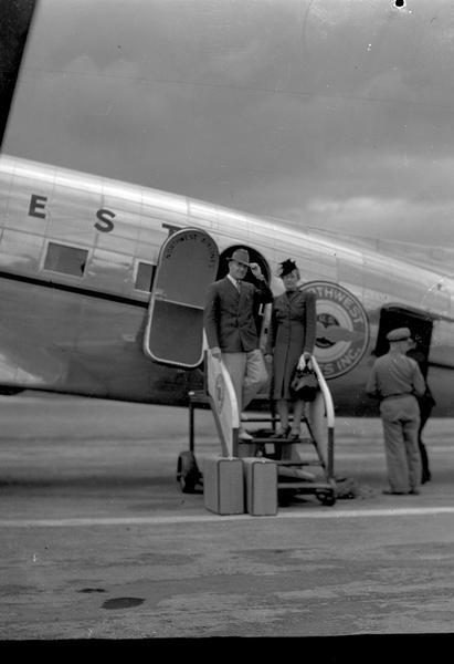 Man and woman with two suitcases boarding a Northwest airplane. "Hundred to One" It's for You" Manchesters Fall merchandise sale. Advertisement says "Now thet you've earned a grand vacation, you'll want to take advantage of the savings on fine luggage."