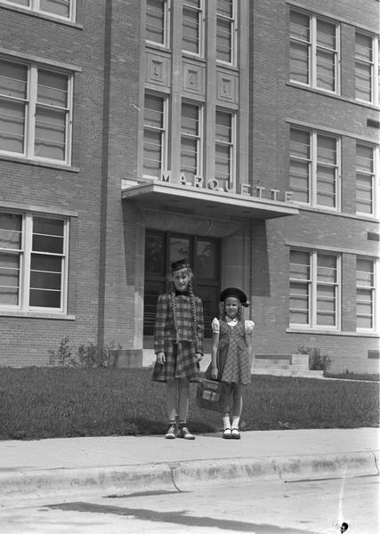 Two young girls standing on the sidewalk in front of the school.