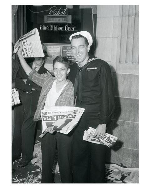 Seaman 1st class Tom Teeley with newsboy, Bernard Ehrmann, holding a copy of the Wisconsin State Journal with headline declaring peace in the Pacific. Celebrating V-J Day, August 15, 1945, the day on which the Allies announced the surrender of Japanese forces during World War II.