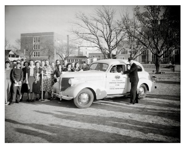 AAA driver's training instructor with group of students, posing with dual control driver's education car, in front of Madison East High School.