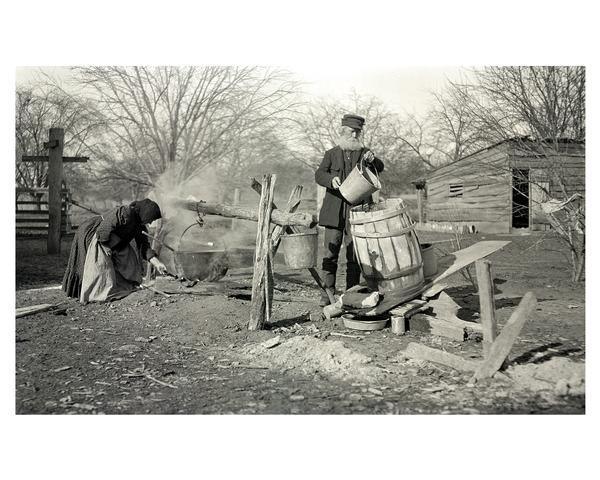 The farmer and his wife are making soap outdoors. Lye is made by letting rain water seep through wood ashes for several months. Lye and fat produce soap. The leach barrel or hopper in this image contains lye.