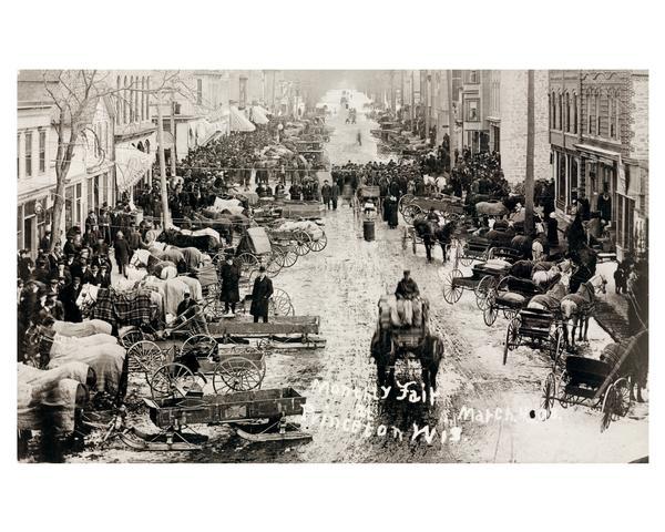 Elevated view of farmer's wagons, teams, and pedestrian traffic crowd Princeton's main street for the monthly market fair. Some snow is on the ground and a few of the wagons have been mounted on runners. All of the horses are covered with blankets to keep them warm. Because horses were crucial to a farmer's livelihood, they required good care.