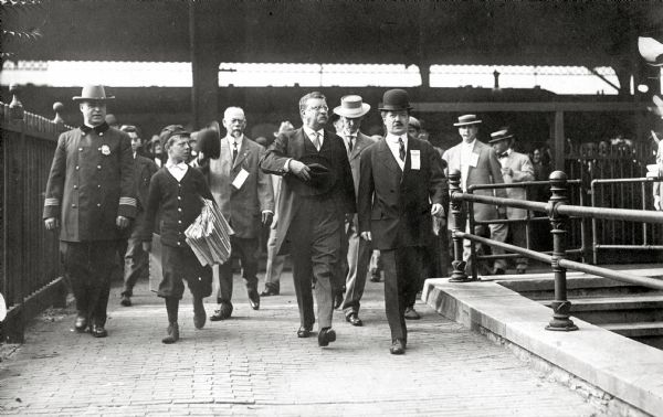 President Theodore Roosevelt leaving the Chicago and North Western depot after arriving in Milwaukee on his October 14th, 1912 visit, during which he was shot and wounded on N. Third Street between W. Wells Street and W. Kilbourn Avenue. Left to right, Police Sgt. Robert Flood; newsboy Howard Cunningham; former Wisconsin Governor George W. Peck; Roosevelt; Oliver Remey, Press Club representative; and Frank Cannon of Milwaukee, then Secretary of the Citizen's Business League and later known as one of the fathers of Wisconsin's modern highway system.