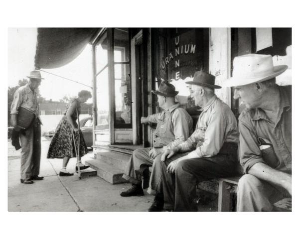 Woman on crutches entering the front office of the uranium tunnel to receive a uranium treatment. Several men sitting in front of the storefront on benches are looking on.