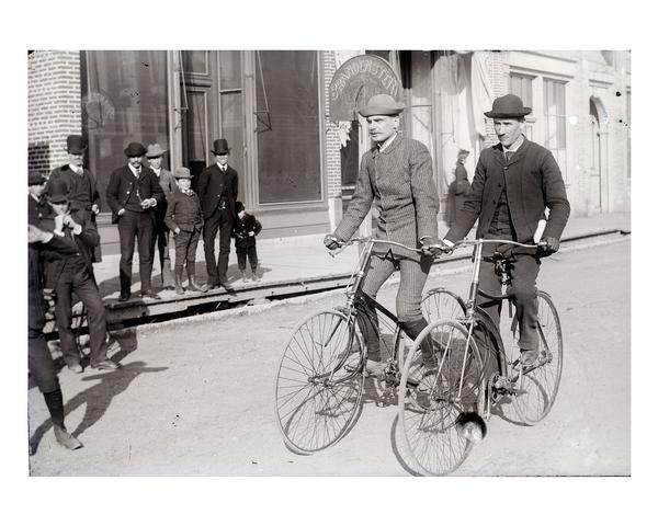 Two men ride their bicycles past curious on-lookers.