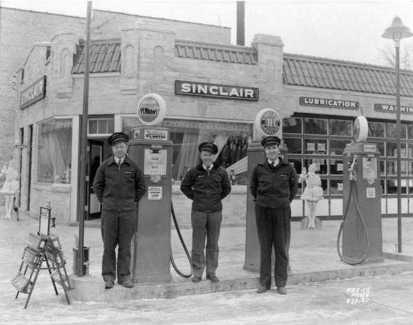 Three uniformed employees of the Sinclair Oil Company Service Station, located at 501 East Broadway, standing in front of the station's gas pumps. Two lifesize cutouts of women are in the background. The station was constructed in 1938.
