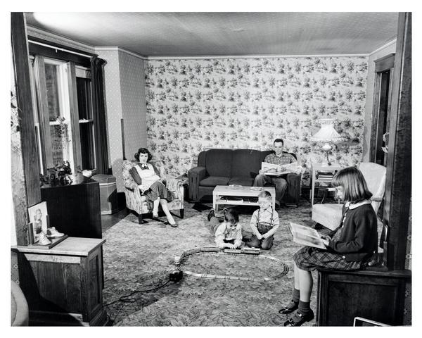 Family in their living room. The mother watches television, the father reads a newspaper, the daughter reads a book, and two sons play with a train set.