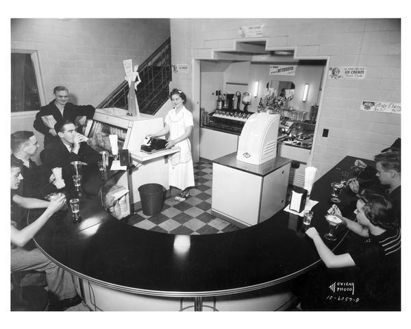Slightly elevated view of a waitress standing behind semi-circular counter with six customers enjoying dairy treats at the Bancroft Dairy Fountain Room, 1010 South Park Street.
