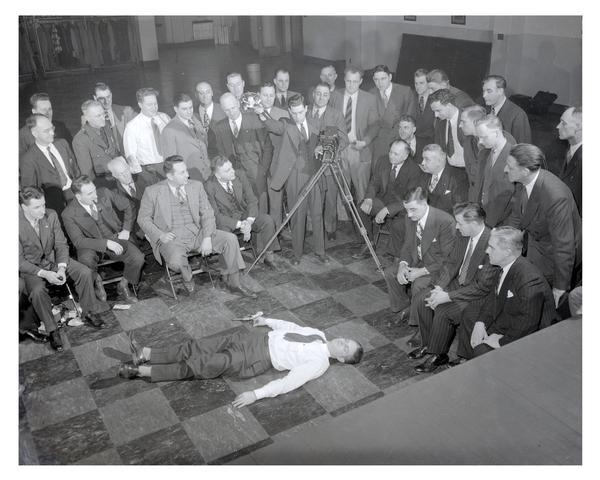 Elevated view of photographers being instructed on how to shoot a crime scene. A teacher is demonstrating photographic techniques while a group of students are observing.