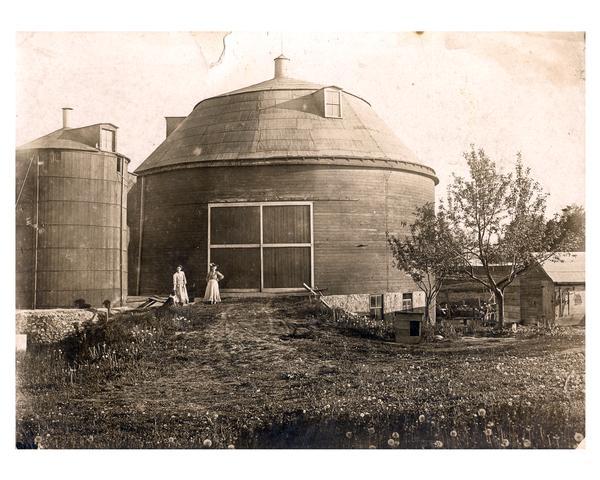 Two women on a farmstead pose in front of a round barn. The name Ri Clausing is written on the back of the photograph.