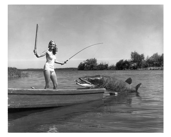 Bathing suit model posing standing in a rowboat, landing an eight-foot plastic Musky (Muskellunge).