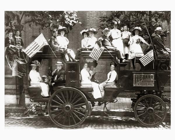 A coaching party of festively clad men, women, and children departing for the Annual Scottish Games sponsored by the St. Andrews Society. Some participants are wearing kilts and tartan sashes. Carriage displays American flags and a sign which reads Hebard, Winchester & Ogden Avenues.