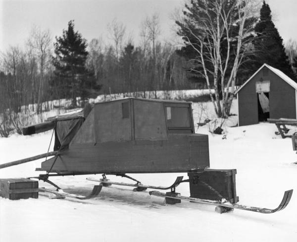 Enclosed wooden box windsled mounted on three skis, sitting on lake ice near the shore.
