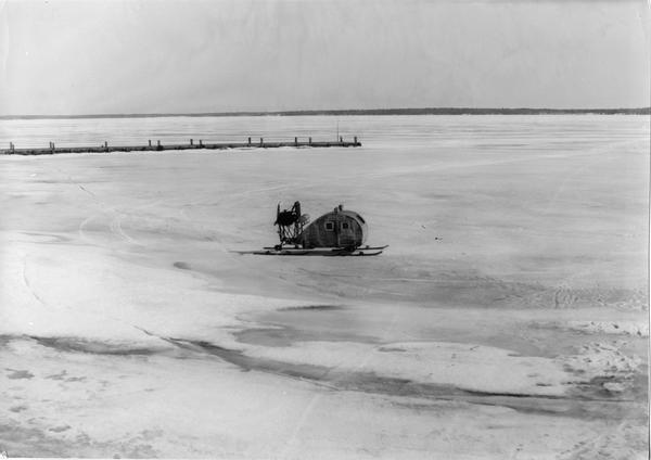 Elevated view of the enclosed wind sled shaped like the <i>Maybe</i>, sitting on the ice with La Pointe dock on the left.