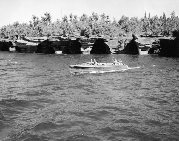 Slightly elevated view of a wooden in-board motorboat on Lake Superior. Two men are in the front seat, and two bathing beauties are riding in the back waving to the photographer. Devils Island sea caves are in the background.