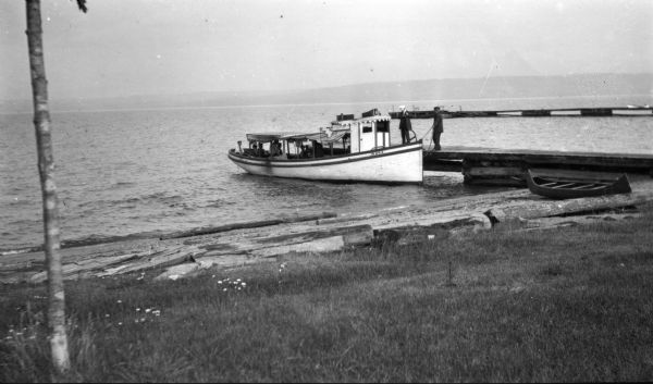View from shoreline of the steamboat "Idora," owned by Charles Russell, docked at the Mission 'stub dock' on Madeline Island. A group of people are on board as two men cast the boat from the dock. A wooden canoe is on shore resting on top of logs. The longer mission dock is in the background, and the far shoreline is in the far background.