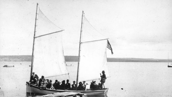 Group of people on sailboat "Alpha" at dock on Madeline Island. Captain Daniel Angus is at the tiller.