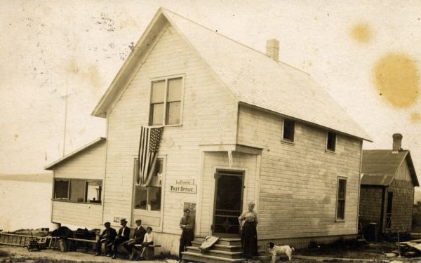 Elizabeth Lathrop, Post Mistress, stands with a dog and other people in front of the Post Office. There is a shoreline on the left.