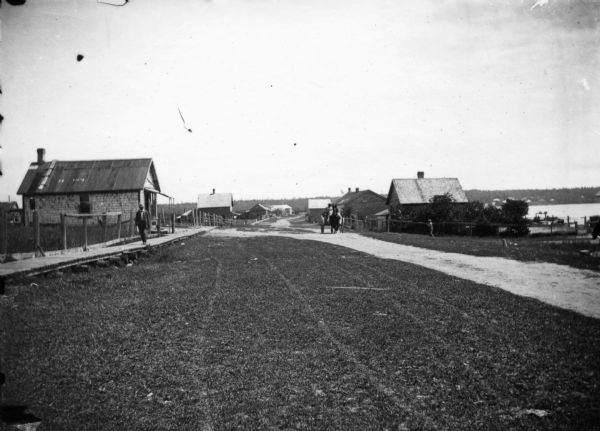 Looking down La Pointe's main street on Madeline Island. A man is walking on the board sidewalk on the left, and another person is driving a horse-drawn vehicle up the unpaved road on the right. The shoreline is in the background on the right.