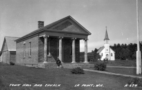 Photographic postcard view of a man sitting on the steps of Town Hall. The Catholic church is in the background. Caption reads: "Town Hall and Church, La Point, Wis."
