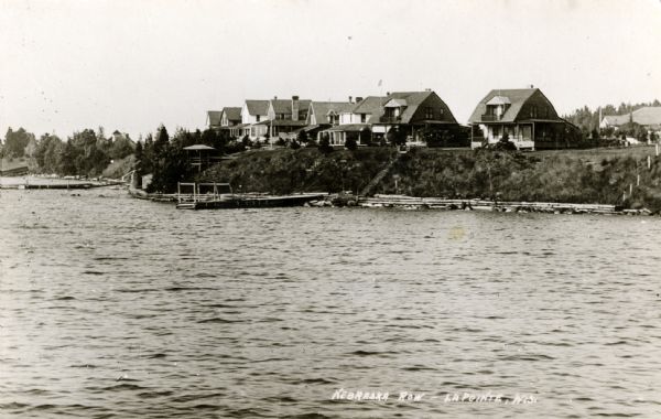 View from water of cottages along the shoreline in Nebraska Row, La Pointe, Madeline Island. Caption reads: "Nebraska Row — La Pointe, Wis."
