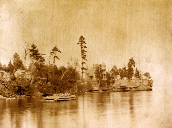 View from Lake Superior of Cedar Bark Lodge and other dwellings on Hermit Island, which has a rocky shoreline. In the water in the foreground is a log-constructed breakwater.