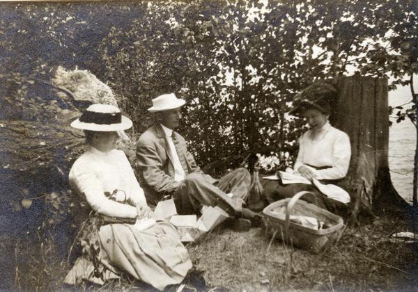 Two women and a man sitting on the shore of Lake Superior, resting against a tree stump and boulders. One woman is reading, with a picnic basket at her side.