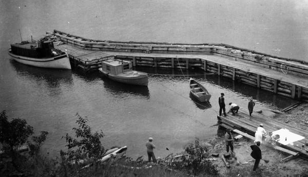 Elevated view of wooden boats at Raspberry Island (or Devils Island) dock with picnickers on shore. The "Lizzie W' and Nelson Angus, A.G. Hull, Elsie Tough, Anne Ashley, Mrs. Hull, Mrs. Baker, Mrs. J.L. Abernathy, Mr. Baker, and Captain Angus. This the dock for the light station.