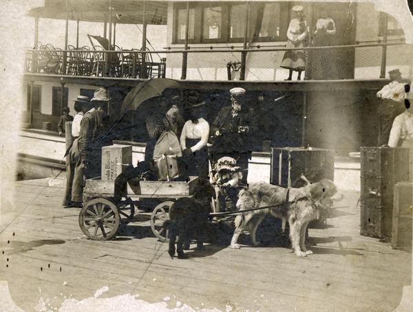Colonel and Mrs. Woods (right center) arriving on Madeline Island dock for summer visit. Thomas Stahl and his dog team (at left), loading luggage into dog cart. Steamer "Plowboy" is in the background.
