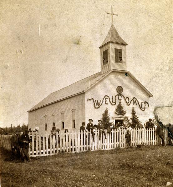 People gathered around picket fence in front of a decorated St. Joseph's Catholic Church in La Pointe on Madeline Island.
