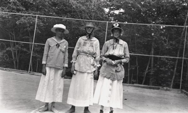 Three young men dressed in women's clothing stand on a tennis court during 4th of July celebration. William Baker, Ted Dwyer, and Herbert Rogers.

