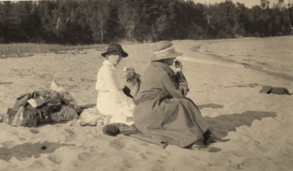 Mrs. Cora Hull & Mrs. Julia Wood are seated on the sandy shore of one of the Apostle Islands overlooking Lake Superior.