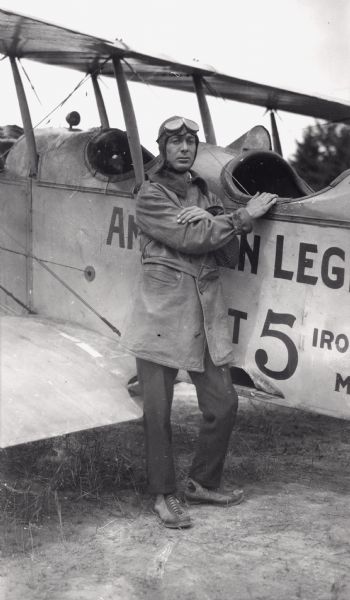 Clarence Russell leaning next to a Jenny bi-plane, wearing aviator gear. This was the first airplane to land on Madeline Island.
