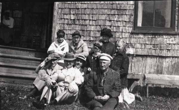 Group of men, women, and children seated on a bench and on the ground outside the Hermitage (Cedar Bark Lodge) on Hermit Island (Wilson Island). Included are Jesse Stone, A.G. Hull, Leo Capser, Captain Angus, Helen Glover, Margaret Glover, Elizabeth Hull, Mr. J.L. Abernathy, and Elizabeth Baker.