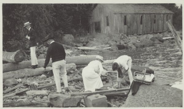 View looking towards shoreline of three men and one woman setting up a fire on the rocks for a picnic. There is a picnic basket on the log on the right, and in the background is a building among the trees.