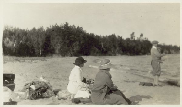 View of Mrs. Cora Hull and Mrs. Julia Wood sitting on the beach at a shoreline. A man stands near the water. In the background are trees.