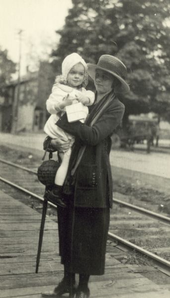 Full-length portrait of Elizabeth Baker standing and holding a young girl in her arms. They are next to railroad tracks.