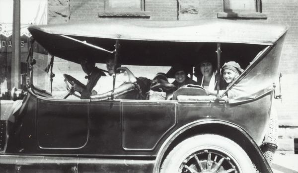Group in automobile looking out the left side windows. A man is driving, a young boy is in the passenger seat, and in the back seat are two women and a young girl. Buildings are in the background.