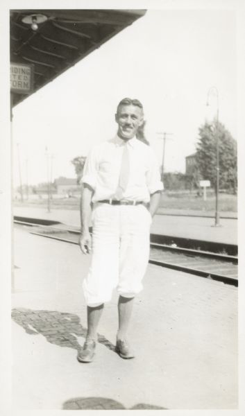 Full-length portrait of Leo Capser standing outdoors in front of railroad tracks. The station roof is on the left.