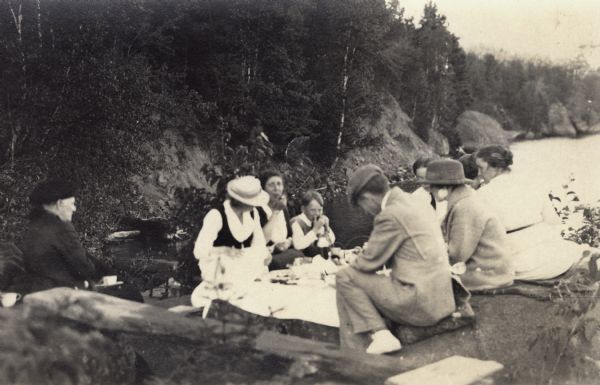 Group of people gathered for a rustic picnic on Bear Island, Apostle Islands, Lake Superior. Image includes Mrs. J.L. Abernathy, Cora A. Hull, Mrs F.B. Chute, John Webb, Rubi Webb, Mrs. Rubi Webb, Mrs. James Austin and Mr. F.B. Chute.
