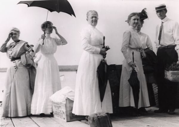 Five ladies and one man standing on Old Mission Dock holding picnic baskets and umbrellas. The ladies with umbrellas are from left: Mrs. Ross, Mrs. Ervine, and Sally Ervine Reitler.  Gene Reitler is holding the basket.