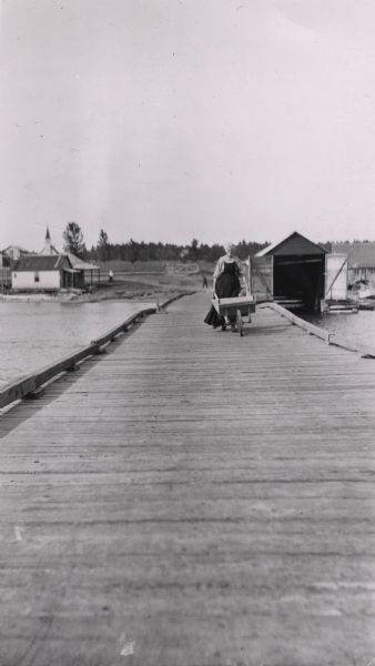 Mrs. Lathrop, the town post mistress, pushing her wheelbarrow down a long wooden dock at La Pointe. She is on her way to haul merchandise for her store.