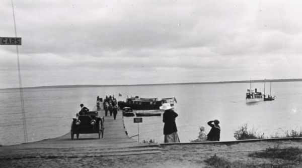 View towards shoreline of an automobile being driven on the Bayfield City Dock. Colonel Woods' boat, "Nebraska" is at the dock. Sam Fifields' "Stella" is at the right near the sailboat. People are on the dock and sitting on the shoreline.