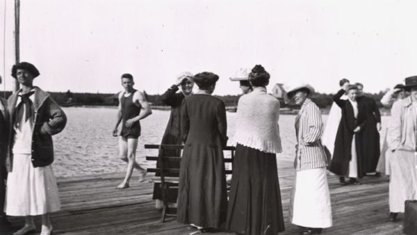 Group of ladies on Mission Dock, and "Ollie" Wilson in bathing suit walking by. Ladies identified as Mrs. N.J. Ross (shielding eyes) and Loretta Salmon in the striped blazer.