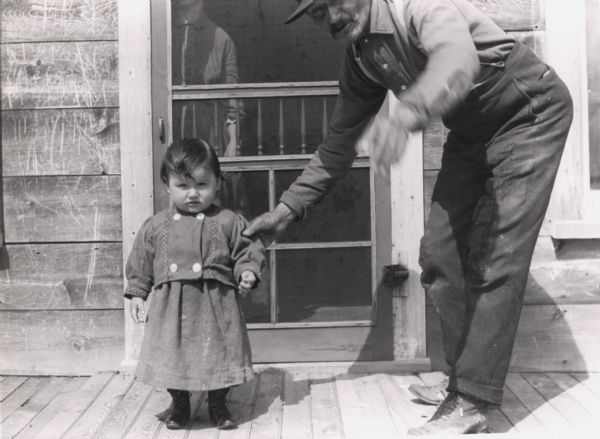 Jean Baptiste "Johnny" Cadotte as a young child on porch on Madeline Island, with unidentified man. A woman is standing inside the screendoor behind them.