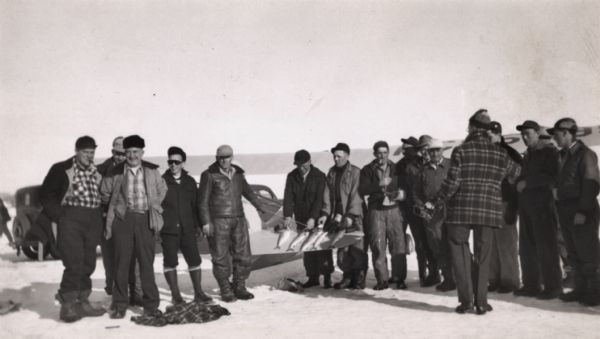 Large group of fishermen standing on ice with catch of fish displayed on airplane wing near La Pointe, Madeline Island.